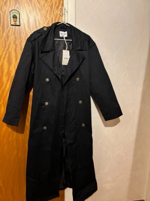 Svart oversized trenchcoat från NA-KD - Stilren svart oversized trenchcoat från NA-KD med dubbelknäppning och klassisk krage. Lång modell med axelklaffar och knappar i metall. Perfekt för en trendig och tidlös look. Inte använd bara provad. 