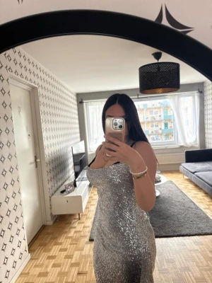Glittrig silver långklänning - Säljer en glittrig silverfärgad långklänning med öppen rygg och smala axelband. Klänningen har en figurnära passform och faller snyggt längs kroppen. Perfekt för dig som vill sticka ut på nästa fest eller bal. 36 och 38 funkar 
