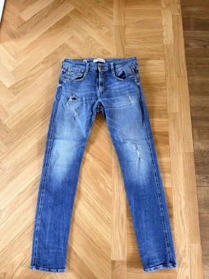 Blå slitna jeans från Replay  - Säljer ett par blå jeans från Replay med slitningar och distressed detaljer på benen. Klassisk femficksmodell med raka ben och läderpatch bak i midjan. Jeansen har en snygg tvättad look och är tillverkade i bomull.