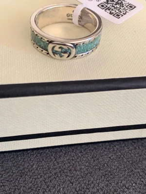 Turkos ring med GG-logo  - Helt oanvänd snygg ring Gucci i silver metall med turkos band runt om och den klassiska GG-logotypen framtill. Ringen har en unik struktur och detaljerade kanter som ger en exklusiv känsla. Perfekt för dig som vill sticka ut med en lyxig  accessoar. Inköpt utomlands lagt ett billigare pris pga ej kvitto el certifikat, inga garantier. Storlek 15. Pris kan diskuteras vid snabb affär.