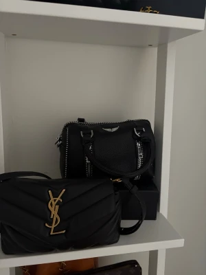 Svart Zadig & Voltaire handväska - Cool svart handväska i läder från Zadig & Voltaire med silverfärgade dragkedjor, nitar och logga framtill. Väskan har både handtag och en justerbar axelrem med broderad VOLTAIRE-text. Perfekt för dig som gillar edgy och trendig stil.