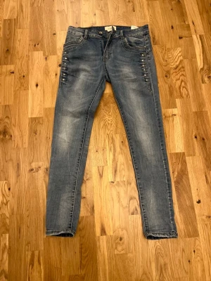 Blå skinny jeans med nitar - Snygga blå skinny jeans med slitningar och coola nitar längs sidorna vid höfterna. Jeansen har klassiska bakfickor och är tillverkade i stretchigt denim för en bekväm passform. Perfekta för dig som gillar en trendig och edgy stil.