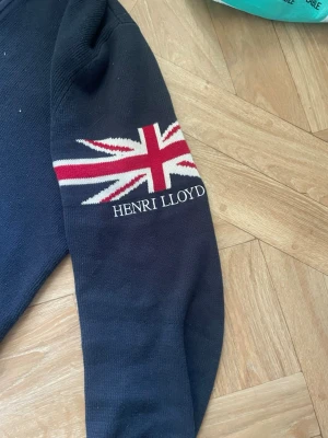 Mörkblå stickad tröja Henri Lloyd - Mörkblå stickad tröja från Henri Lloyd med Union Jack-motiv och logotyp på ena ärmen. Tröjan har ribbstickade muddar och ett broderat emblem på nederkanten. Perfekt för dig som gillar klassisk brittisk stil och vill ha något snyggt och varmt.