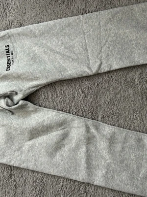 Grå mjukisbyxor Essentials Fear of God - Snygga grå mjukisbyxor från Fear of God Essentials med svart logga tryckt på låret. Byxorna har en klassisk rak passform och är gjorda i mjukt och bekvämt material, perfekt för en avslappnad stil.