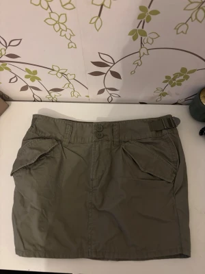 Olivgrön kortkjol med fickor - Snygg olivgrön kortkjol med två stora fickor fram och bak, samt justerbara spännen i midjan. Kjolen har knäppning med två knappar framtill och är perfekt för en avslappnad och cool stil.