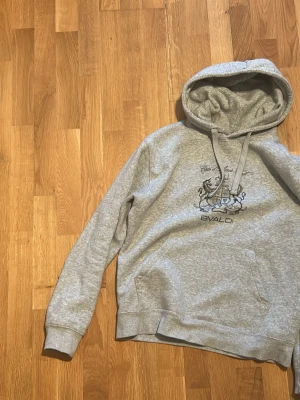 Grå hoodie från Bvaldi med tryck - Säljer en grå hoodie från Bvaldi med svart tryck framtill och klassisk känguruficka. Tröjan har dragsko i huvan och ribbade muddar vid ärmslut och nederkant. Tillverkad i mjukt sweatshirtmaterial, perfekt för en avslappnad stil. Nypris 900kr