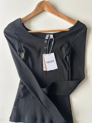 Svart ribbad långärmad topp Pieces - Svart ribbad långärmad topp från Pieces med rund halsringning och knappar framtill. Snygg och enkel modell som är lätt att matcha till olika outfits. Aldrig använd och den är i storlek XS 