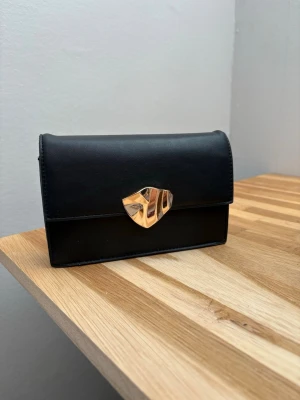 Svart kuvertväska med gulddetalj - Stilren svart kuvertväska/clutch i slätt material med unik guldfärgad låsdetalj framtill. Väskan har en klassisk rektangulär form och justerbara band som går att ta av. Köpt på Kappahl och perfekt att använda på bal/sittning/vanlig utekväll🤗