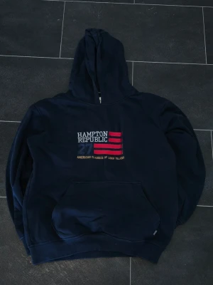 Marinblå hoodie Hampton Republic - Mörkblå hoodie från Hampton Republic med broderad text och detaljer i vitt, rött och gult framtill. Klassisk känguruficka och stor huva. Snygg amerikansk college-stil med trycket '27' och 'American Classics of Long Island'. Perfekt för en avslappnad look.