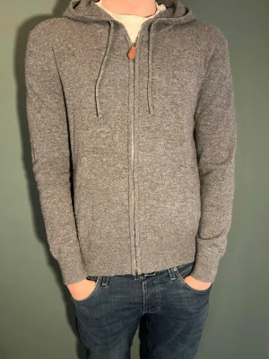 Grå zip hoodie i mjuk ullmix - Säljer en stilren grå hoodie med dragkedja och huva. Tröjan har dragsko i huvan och ribbade muddar vid ärmslut och nederkant. Tillverkad i en mjuk ullblandning som känns riktigt skön mot huden. Perfekt för dig som gillar en enkel men snygg look. Nypris ca 1100 kr
