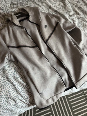 Grå Nike Tech Fleece hoodie - Snygg grå Nike Tech Fleece hoodie med dragkedja och svarta detaljer längs bröstet. Klassisk huva och två sidofickor. Perfekt för en sportig och avslappnad stil. Materialet är mjukt och stretchigt för extra komfort.
