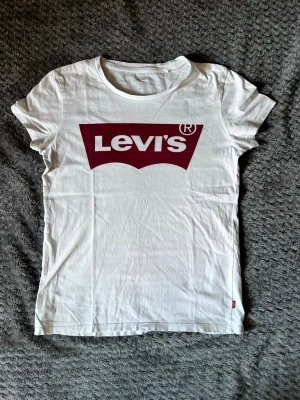 Vit Levi's t-shirt med röd logga - Vit t-shirt från Levi's med klassisk röd logga framtill. T-shirten har rund halsringning och korta ärmar. Tillverkad i mjuk bomull, perfekt för en avslappnad och stilren look.