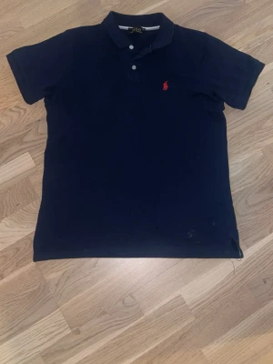Marinblå pikétröja från Polo Ralph Lauren - Klassisk marinblå pikétröja från Polo Ralph Lauren med korta ärmar och två knappar vid kragen. Tröjan har den ikoniska röda logotypen broderad på bröstet och är tillverkad i mjuk bomull. Perfekt till jeans eller shorts.
