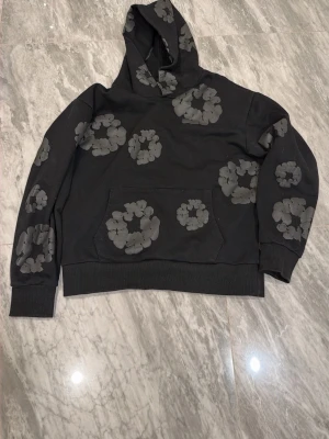 Svart hoodie från Denim Tears - Svart hoodie från Denim Tears med unika gråa blomliknande tryck över hela plagget. Hoodien har en stor huva och en klassisk känguruficka framtill. Perfekt för dig som vill sticka ut med en streetwear-look.