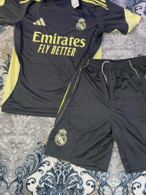 Real Madrid träningsset Adidas svart/gul - Snyggt träningsset från Real Madrid med svart t-shirt och shorts, båda med gula detaljer och klubbens logga. Tröjan har trycket 'Emirates Fly Better' och Adidas-logga. Perfekt för fotbollsträning eller gym. Materialet är lätt och andas.