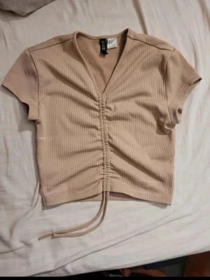 Beige ribbad croppad topp - Trendig beige croppad topp med ribbad struktur och snygg v-ringning. Toppen har korta ärmar och snörning framtill som ger en cool detalj. Perfekt att styla med jeans eller kjol. Liten missfärgning på sidan av magen men går säkert bort i tvätt. 