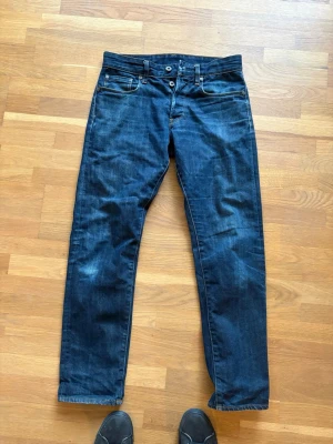 Mörkblå raka jeans från g star raw - Snygga mörkblå jeans från Nudie med klassisk rak passform och fem fickor. Jeansen har en enkel design med kontrastsömmar och läderpatch bak i midjan. Perfekta för en avslappnad och stilren look.