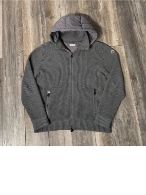 Moncler cardigan - Snygg grå cardiganjacka från Moncler med stickade ärmar och framstycke samt quiltad rygg och huva i nylon. Jackan har dragkedja framtill, två fickor med dragkedja och Moncler-logga på ärmen. Perfekt för lager på lager och streetwear. Passar dig som har L. Behöver pengar snabbt så där av priset