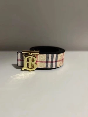 Rutig bälte med gulddetalj - Stilrent bälte med klassiskt rutigt mönster i beige, rött och svart. Bältet har ett exklusivt spänne i guld med B-logga. Perfekt accessoar för att lyfta din outfit.