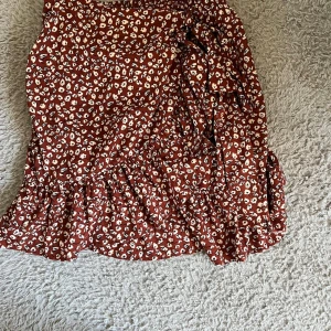 Blommig brun omlotkjol med volang - Supersöt brun omlottkjol med vitt blommönster och volangkant. Den  har knytband  och en luftig passform som ger en avslappnad vibe. Perfekt för dig som vill ha en trendig och bekväm look till sommaren.