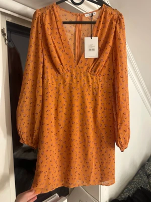 Orange blommig klänning från NA-KD - Kort klänning från NA-KD i orange med små rosa blommor. Djup V-ringning och långa puffiga ärmar.