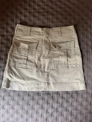 Beige kortkjol från Gina Tricot - Snygg beige kortkjol från Gina Tricot med två stora fickor bak och bälteshällor. Kjolen har en rak passform och är något liten i storleken