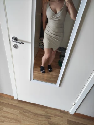 Beige kortklänning med v-ringning - Snygg beige kortklänning med omlott och djup v-ringning. Klänningen är ärmlös och har en figurnära passform som framhäver siluetten. Perfekt för dig som gillar stilrena och trendiga plagg. Köppt på modebutik i Spanien. 