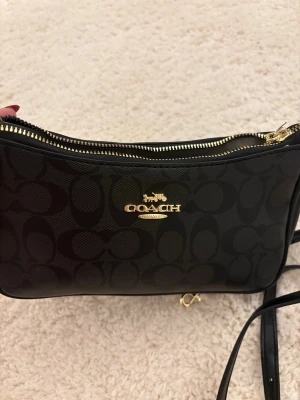 Svart handväska från Coach - Snygg svart handväska från Coach med klassiskt monogrammönster och guldfärgade detaljer. Väskan har dragkedja upptill och justerbar axelrem. Perfekt för att bära det viktigaste och ger en lyxig känsla till din outfit.