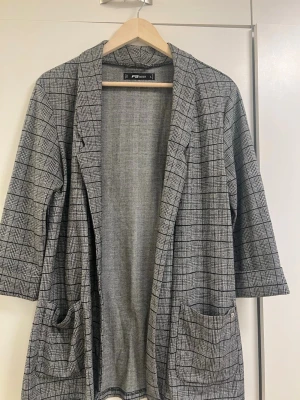 Grå rutig kavaj från FB Sister - Snygg grå kavaj från FB Sister med svartvitt rutmönster. Klassisk krage, fickor framtill och rak passform. Perfekt för en trendig och stilren look. Materialet känns mjukt och lätt, vilket gör den bekväm att bära hela dagen.