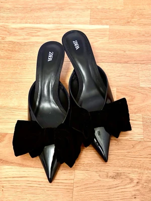 Svarta klackar med rosett från Zara - Säljer ett par svarta pumps från Zara med spetsig tå och låg klack. Skorna har en stor sammetsrosett framtill som ger en extra stylish touch. Tillverkade i glansigt material och är öppna baktill.