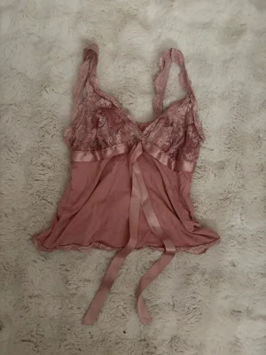 Rosa spetsbody med rosett - Söt rosa body med spetsdetaljer upptill och breda axelband. Satinband under bysten som knyts till en rosett framtill. Tunt, mjukt material som ger en romantisk känsla. Perfekt för dig som gillar feminina och eleganta underkläder.