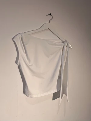Vit oneshoulder topp med knyt - Snygg vit topp i oneshoulder-modell med knyt på axeln. Toppens enkla design gör den lätt att matcha och den är tillverkad i ett mjukt, stretchigt material som sitter bekvämt. Perfekt för dig som gillar stilrena och trendiga plagg.