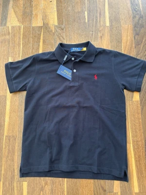 Mörkblå pikétröja  Polo Ralph Lauren - Klassisk mörkblå pikétröja från Polo Ralph Lauren med röd broderad logga på bröstet. Tröjan har korta ärmar, krage och två knappar framtill. Tillverkad i mjuk bomull som känns skön mot huden. Perfekt för en stilren och avslappnad look.