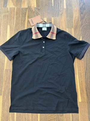 Svart pikétröja Burberry - Svart pikétröja från Burberry med klassisk krage i beige och rutig design med röda detaljer. Tröjan har korta ärmar och knäppning framtill. Perfekt för en stilren och trendig look.