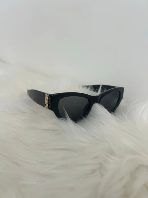 Saint Laurent sol glasögon  - Snygga svarta solglasögon i cat eye-modell med cool guldfärgad detalj på sidorna. Glasögonen har en stilren och trendig look som passar perfekt till sommaren. Ramen är i plast och glasen är mörka för ett edgy intryck.