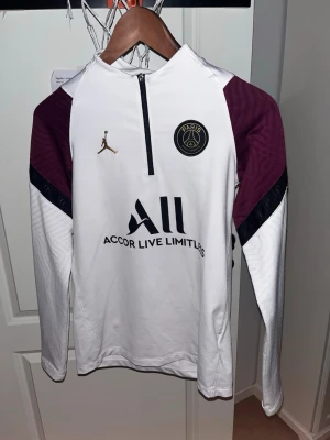 PSG träningströja vit/bordeaux - PRIS KAN DISKUTERAS. L i barn men passar XS. Snygg långärmad träningströja från PSG och Jordan i vitt med bordeauxfärgade axlar och svarta detaljer. Halv dragkedja framtill, klubbmärke och Jordan-logga på bröstet. Perfekt för fotbollsträning eller chill.