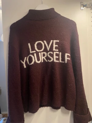 Vinröd stickad tröja med text - Mysig vinröd stickad tröja med polokrage och vit texten 'LOVE YOURSELF' framtill. Tröjan har långa ärmar och ribbade muddar vid hals, ärmslut och nederkant. Perfekt för kalla dagar och ger en skön, avslappnad look.