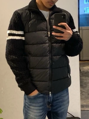 Svart pufferjacka från Moncler - Svart pufferjacka från Moncler med stickade ärmar och två vita ränder på varje ärm. Jackan har dragkedja framtill, ståkrage och två fickor med dragkedja. Ärmarna är ribbstickade och har Moncler-logga på vänster ärm. Perfekt för kalla vinterdagar. INTE ÄKTA!