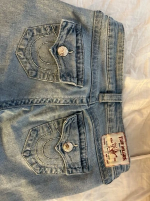 Ljusblå lågmidjade jeans från True Religion - Säljer ett par ljusblå jeans från True Religion med ikoniska bakfickor med lock och silverfärgade knappar. Jeansen har klassisk femficksdesign och patch med logga bak i midjan. Perfekta för dig som gillar street och premium denim. storlek 25, använt några gånger så dom är lite slitna på sömmen längst ner på byxorna. 
