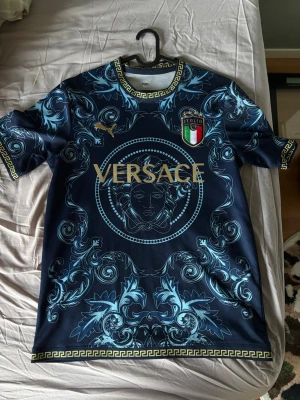 Blå V****e fotbollströja Italien - Snygg blå fotbollströja från V*****e med guldiga detaljer och Medusa-motiv. Tröjan har Italiens landslagsemblem, guldtexten 'Jorginho' och nummer 8 på ryggen. Unik design med grekiska mönster längs krage och ärmslut. Perfekt för dig som vill sticka ut på planen.