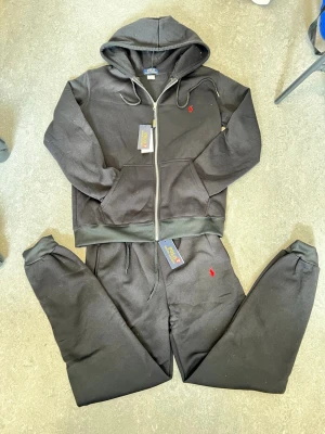 Svart loungewear-set från Polo Ralph Lauren - Mysigt svart loungewear-set från Polo Ralph Lauren med zip-hoodie och matchande byxor. Setet har röda broderade loggor på både tröjan och byxan, dragsko i midjan och mjukt material. Perfekt för chill dagar hemma.
