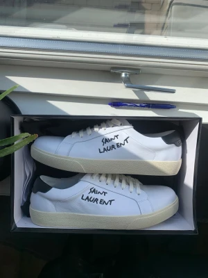 Vita sneakers  - Säljer ett par vita Saint Laurent sneakers med svart häl och handskriven logga på sidan. Skorna har klassisk låg profil, vita snören och sula i offwhite. Tillverkade i läder med snygg minimalistisk design och street-inspirerad vibe. Skriv om du har några funderingar!😁