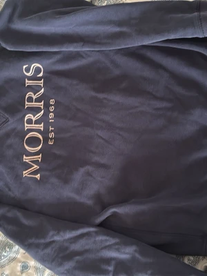 Mörkblå sweatshirt från Morris - Snygg mörkblå sweatshirt från Morris med stort tryck 'MORRIS EST 1968' på framsidan. Tröjan har rund halsringning och ribbade muddar vid ärmslut och nederkant. Perfekt för en avslappnad och stilren look.