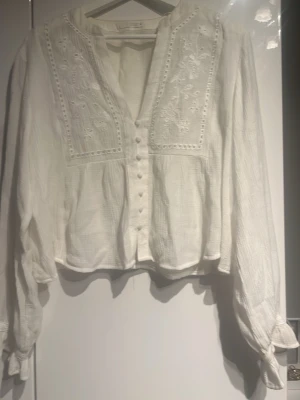 Vit blus med broderi från Zara - Vit blus från Zara med långa ärmar, knappar framtill och vackra broderade detaljer på bröstet. Blusen har en lätt, luftig struktur och en avslappnad passform. Perfekt för en trendig och stilren look.