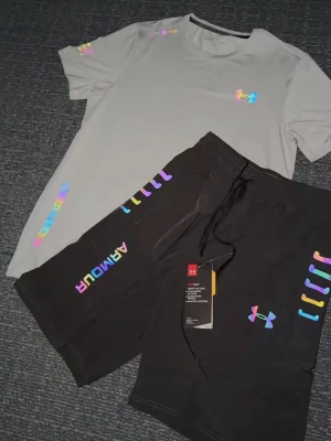 Grå träningsset från under armour - Snyggt set från Under Armour med en ljusgrå t-shirt och svarta shorts. Båda plaggen har reflektiva loggor och textdetaljer som ger en modern vibe. Perfekt för dig som gillar sportig och trendig stil. Materialet känns mjukt och bekvämt mot huden.