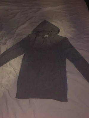 Grå Kashmir hoodie  - En enkel och stilren kashmir hoodie med huva. Hoodien har lång ärm och är tillverkad i ett mjukt, stickat material som känns skönt mot huden. Perfekt för dig som gillar en clean och avslappnad look.