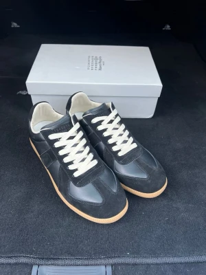 Svarta sneakers från Maison Margiela - Säljer ett par svarta sneakers från Maison Margiela med klassisk design. Skorna har ovandel i svart läder och mocka, vita snören och beige gummisula. Insidan är vit och modellen är stilren och tidlös, perfekt för dig som gillar minimalistisk streetwear.
