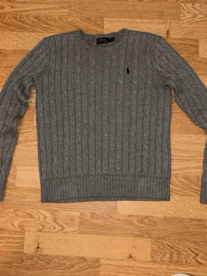 Grå kabelstickad tröja Polo Ralph Lauren - Snygg grå kabelstickad tröja från Polo Ralph Lauren med rund halsringning och broderad logga på bröstet. Tröjan har långa ärmar och ribbade muddar vid ärmslut och nederkant. Perfekt för lager på lager och klassisk stil.