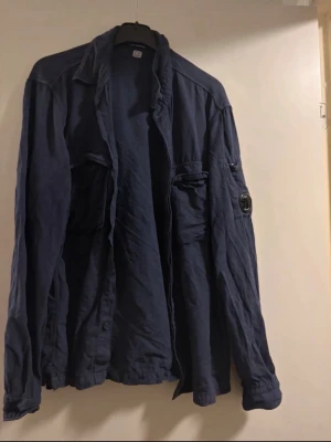 Mörkblå overshirt från C.P. Company - Snygg mörkblå overshirt från C.P. Company med klassisk krage, knappstängning och flera fickor framtill. Ärmen har det ikoniska linsmärket. Tillverkad i bomull för en skön och avslappnad känsla. Perfekt för lager-på-lager-stil.