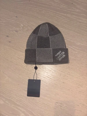 Grå rutig mössa - Säljer en snygg grå mössa  med rutigt mönster och uppvikt kant. Mössan har broderad text på framsidan och är tillverkad i ett mjukt stickat material som passar perfekt för kyliga dagar.